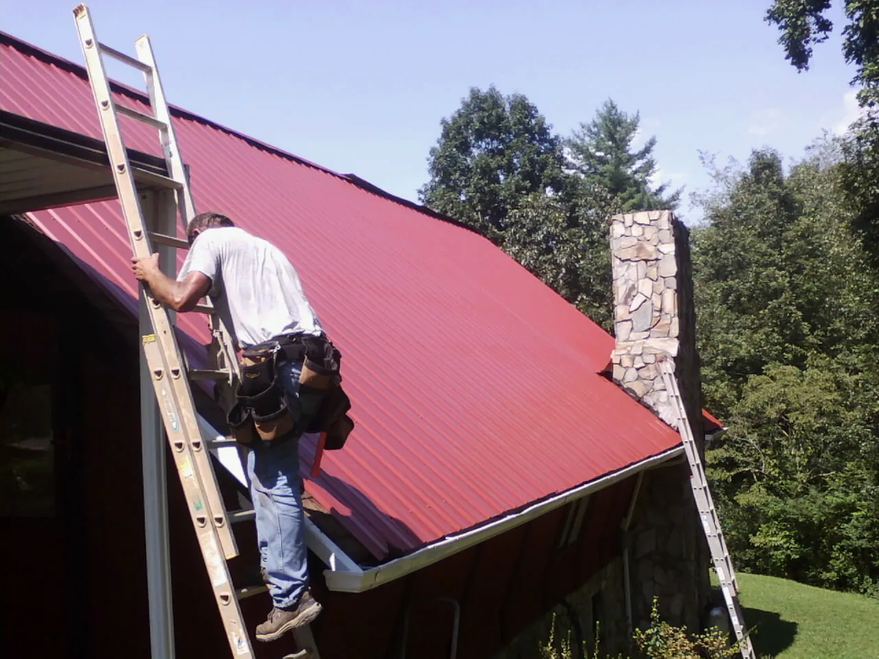 Roofer on a ladder beside a red metal roof and stone chimney — Soffit & Fascia Repair by Roofing Brothers in Farragut